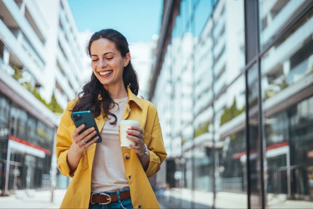 Smiling woman standing outdoors in an urban setting, looking at her smartphone while holding a coffee cup.