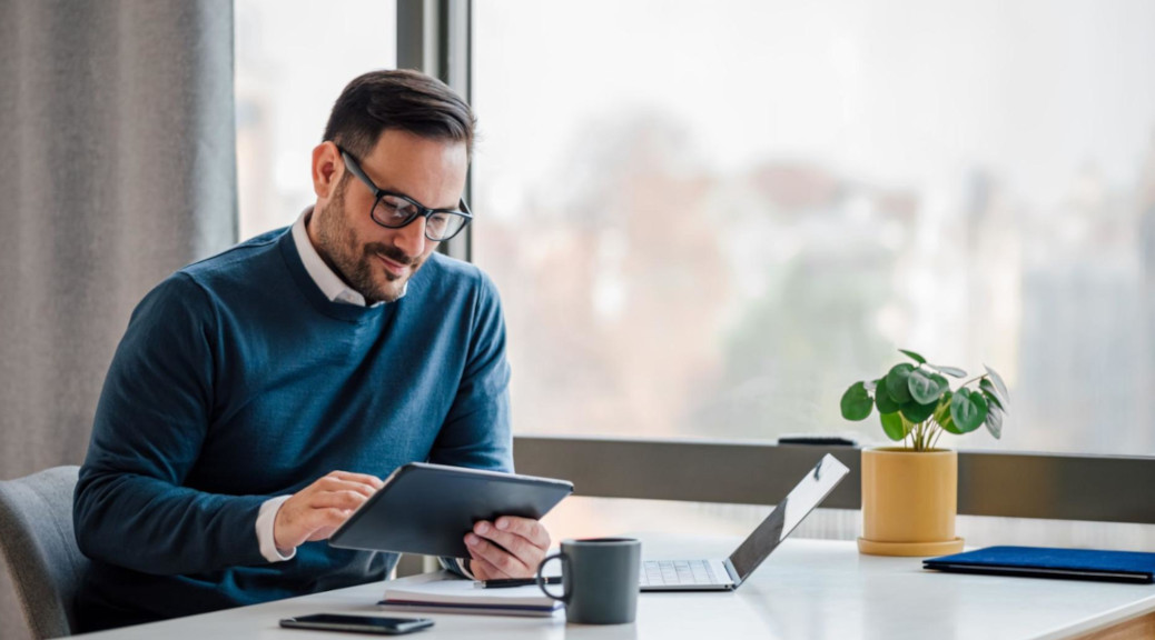 Business professional in a modern office setting, using a digital tablet while sitting at a desk with a laptop.