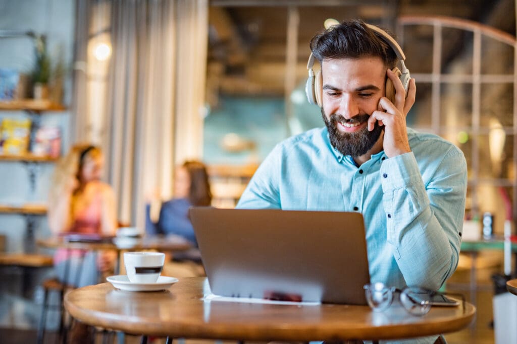 A young professional working remotely in a café, wearing headphones and speaking on a VoIP call while using a laptop.