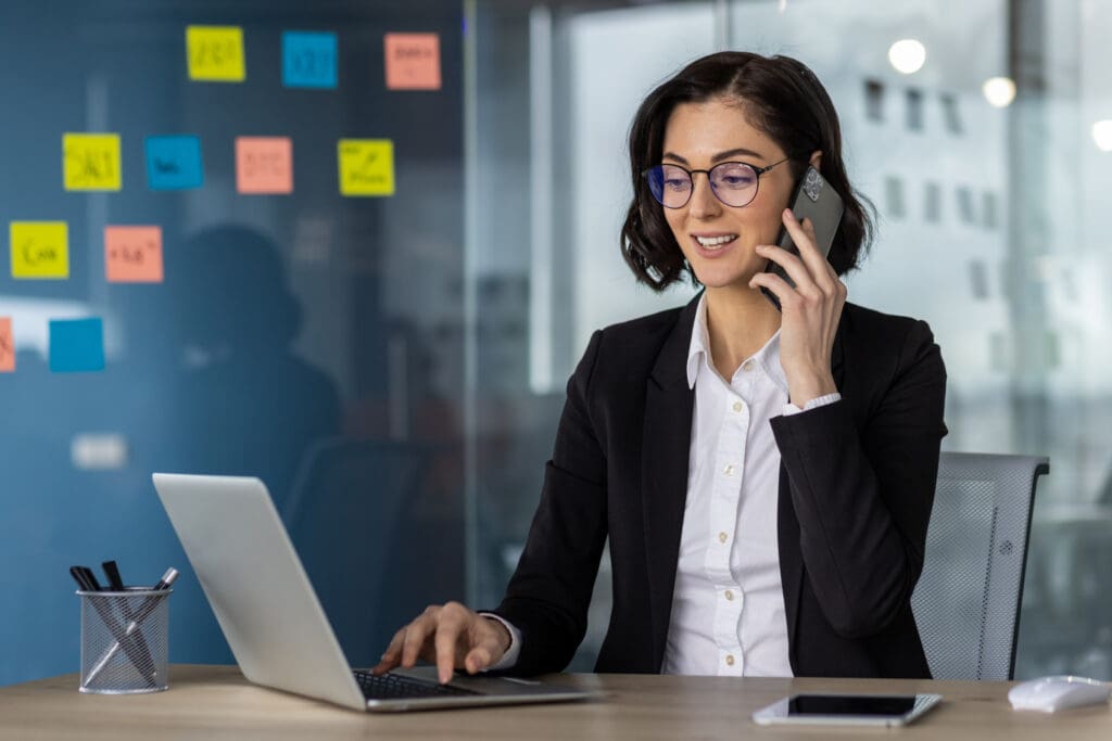 A businesswoman in an office setting speaking on a smartphone while using her laptop.