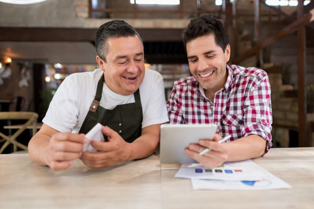 Two small business owners happily reviewing financial documents and discussing savings while using a tablet in a cozy office environment.