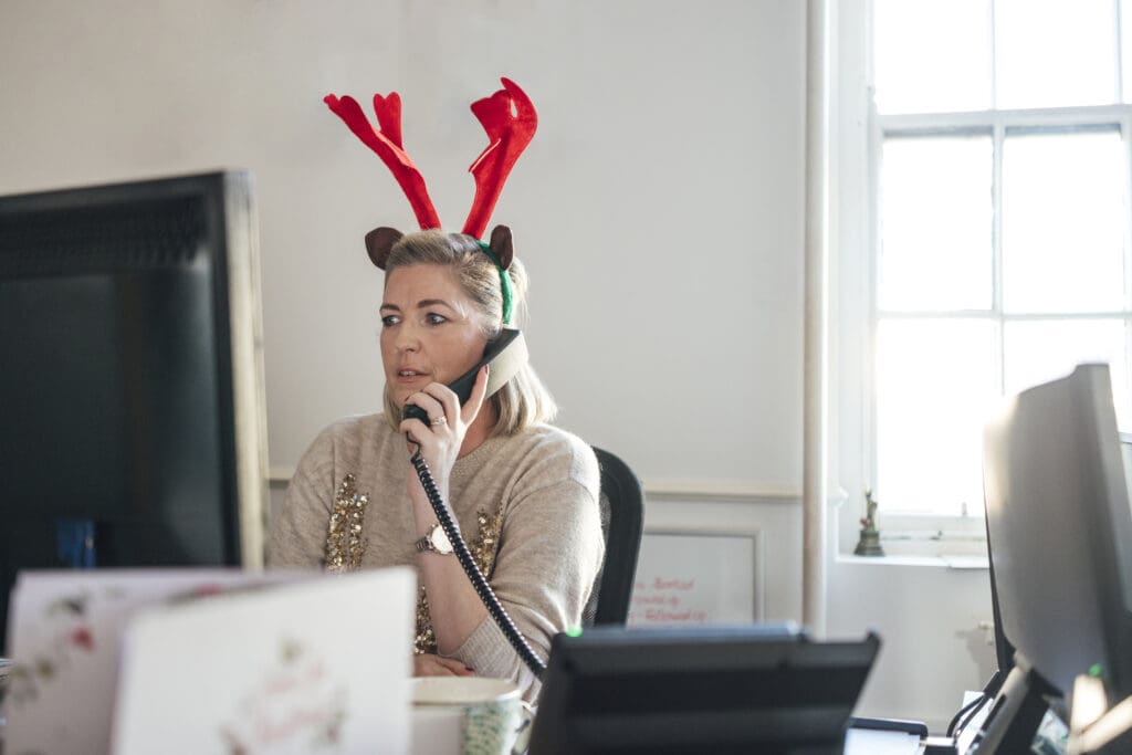 Woman in a festive reindeer headband using a desk phone in an office decorated for the holidays.