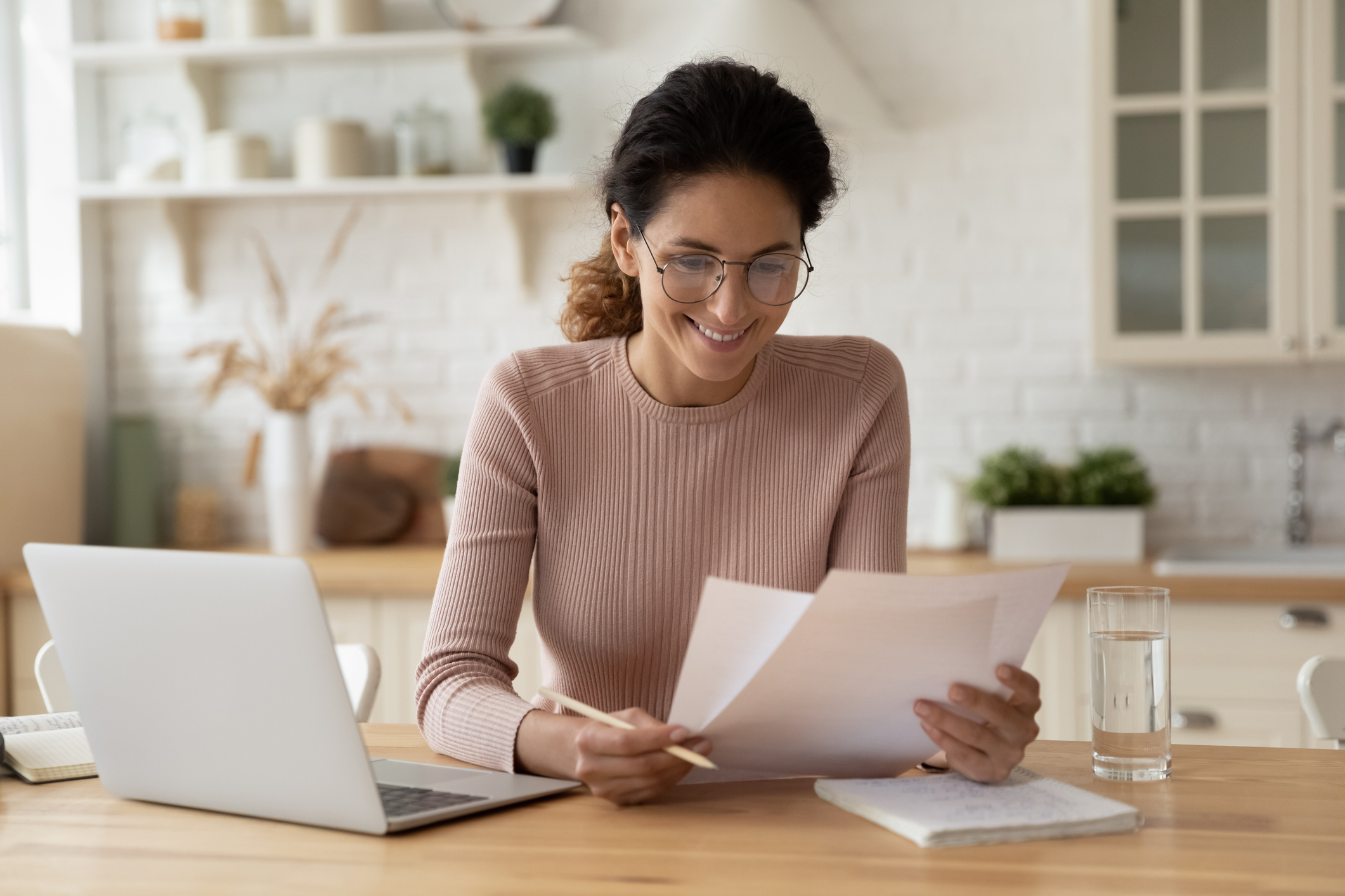 Smiling woman reviewing financial documents at home with a laptop and notepad on the table, representing cost savings and efficiency with VoIP and SMS communication.